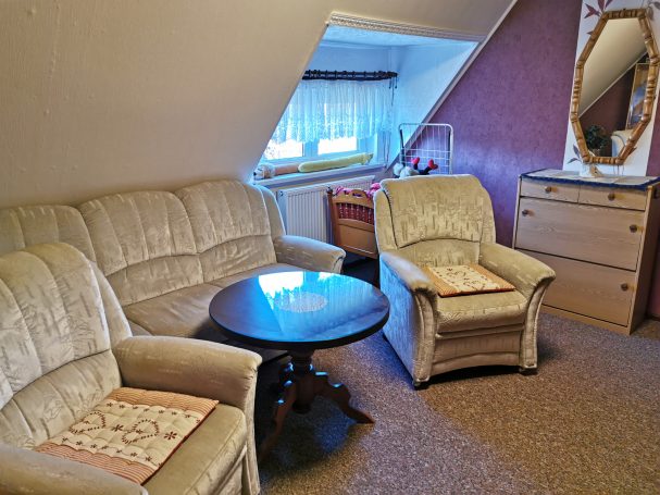 Gemütliches Wohnzimmer mit Sofa, Sessel, Tisch und Kommode. Helle Fenster und dekorative Elemente.