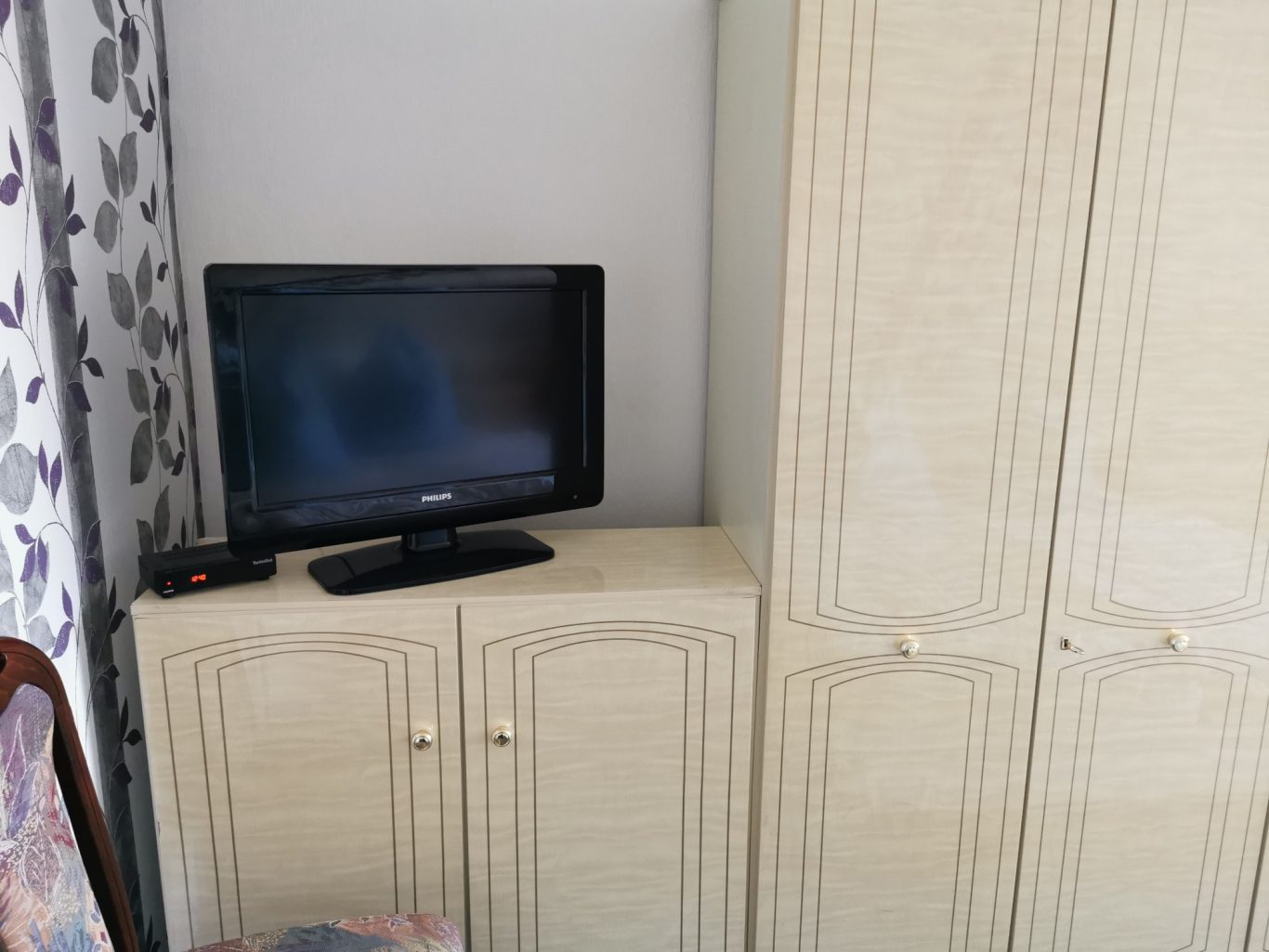 Ein Fernseher auf einem beige Farben Schrank, daneben ein hoher Kleiderschrank.