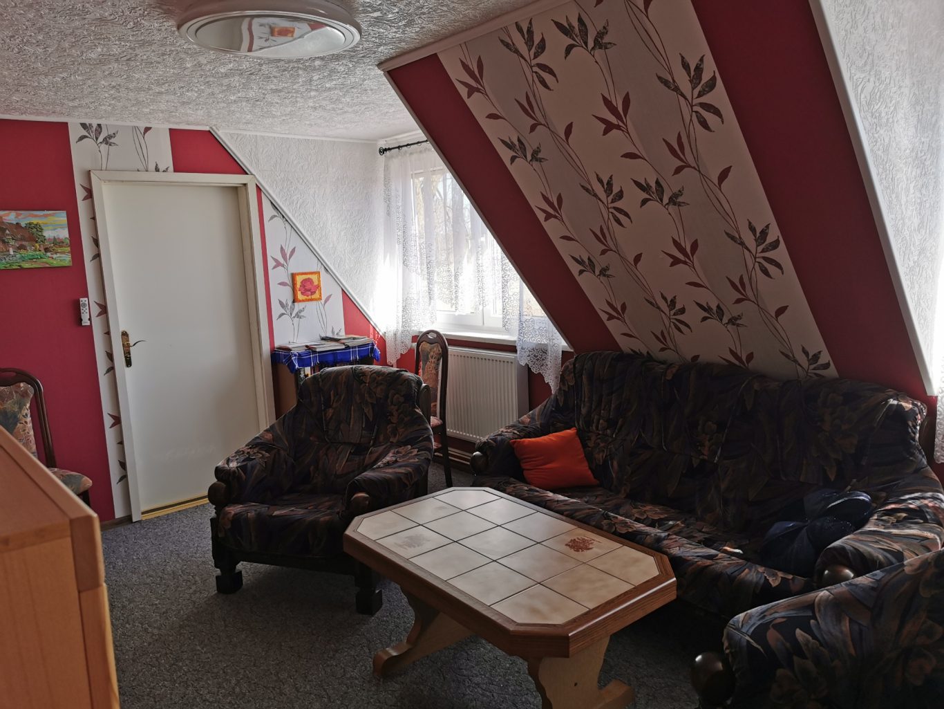 Gemütliches Wohnzimmer mit zwei Sessel, Sofa und Tisch, rote und blühende Wandgestaltung.