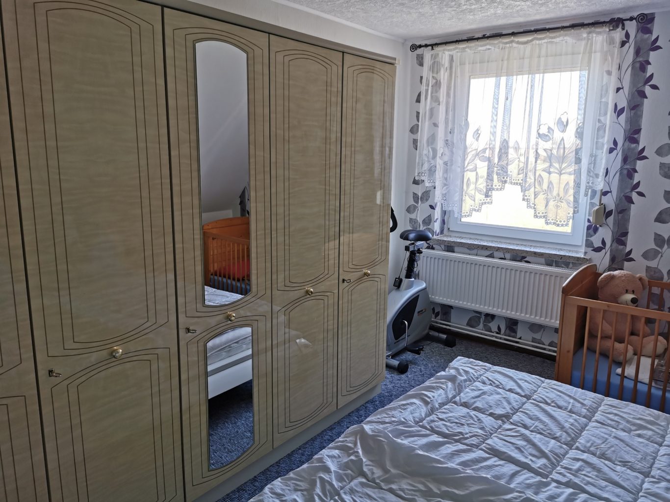 Schlafzimmer mit Schrank, Fenster, Bett und Babybett im Hintergrund.