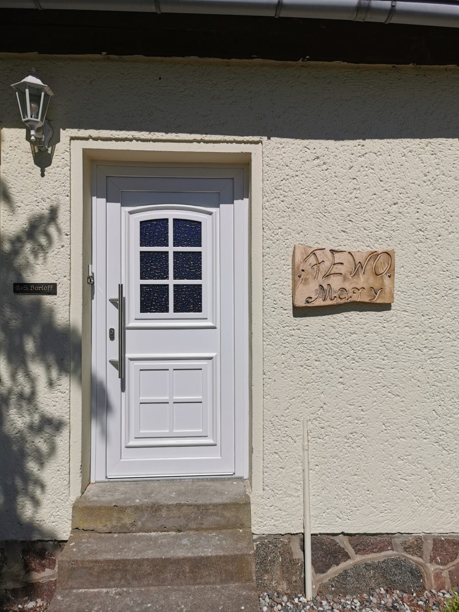 Weiße Tür mit Fenster und Holzschild an einem hellen Gebäude.