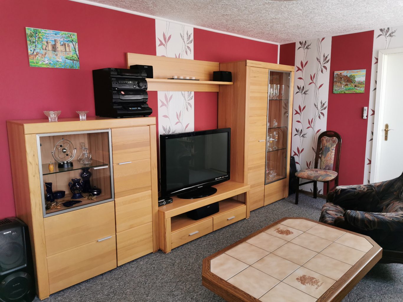 Wohnzimmer mit hellen Möbeln, Fernseher, Regal, Tisch und gemütlichem Sessel.